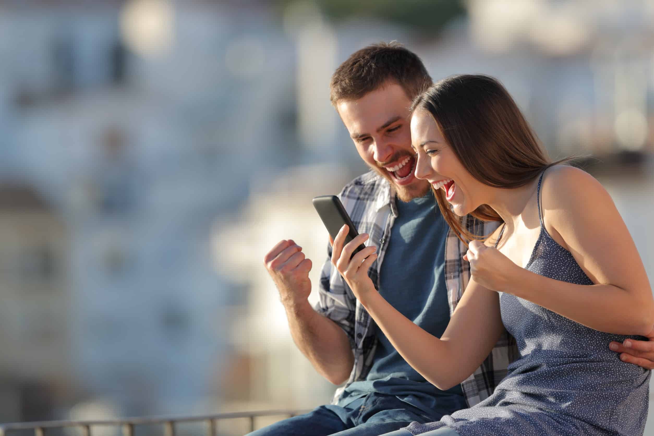 Couple excité célébrant les nouvelles en ligne sur les téléphones intelligents à l'extérieur dans une périphérie de la ville au coucher du soleil